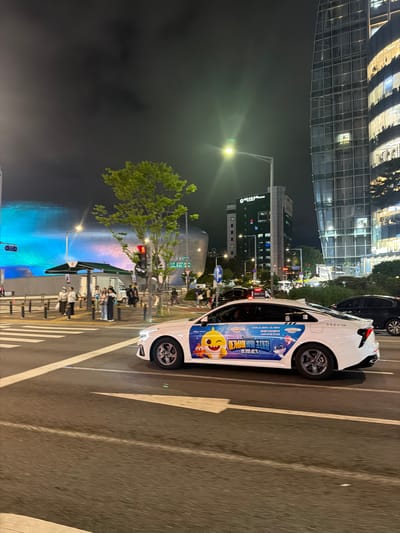 The image depicts a street scene at night in the city of Seoul, South Korea, likely in the vicinity of the Dongdaemun Design Plaza (DDP). On the right side of the frame, a modern, glass-facade skyscraper with illuminated windows dominates the background. In the mid-ground, a large, curved building with blue and pink lighting, characteristic of the DDP, is visible.
In the foreground, a white car, possibly a taxi, is driving on a street. The car is decorated with a large graphic of Baby Shark characters, suggesting a promotional wrap. Several people are visible near a bus stop on the left side of the street, waiting or walking. Traffic lights are present, and the street is marked with crosswalks. The overall atmosphere is urban and illuminated by streetlights and building lights, indicating nighttime.