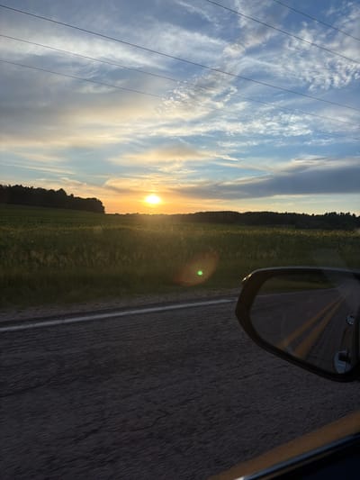 This is a photograph taken from a vehicle, showing a landscape view at sunset. In the foreground, the asphalt road is visible, with yellow lane markings reflected in the car's side mirror. The side mirror itself is also in focus, showing a portion of the road and sky. Beyond the road, a vast green field stretches out, dotted with what appear to be crops or tall grass. A dense line of trees forms the horizon, silhouetted against the vibrant sunset sky. The sky is a mix of blue, orange, and white clouds, with the sun positioned low on the horizon, casting a warm glow over the scene. Power lines are visible traversing the upper portion of the sky. The location is specified as the Town of Dell Prairie, United States of America. There are no people visible in the image.