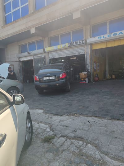 A photograph taken on a sunny day shows the exterior of a building in Baku, Azerbaijan, featuring an auto repair shop. The building has a light-colored facade with several windows. Above the shop entrance, the words "QISMET" and "SLESAR" are visible. The shop appears to have two open bays with metal roll-up doors.
In the foreground, a white car is partially visible, parked on a cobblestone and asphalt surface. To the right of the white car, a black Kia sedan with a license plate reading "77-EX-147" is parked, its rear end facing the camera. A man wearing a dark t-shirt is visible near the open hood of a light-colored car parked to the left of the Kia, possibly working on it. Various automotive supplies, including boxes labeled "Petrovoil," are stacked near the shop entrance. The overall scene depicts a busy auto repair service with cars and workers present.