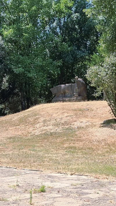 The image shows a monument of a wolf suckling two children, possibly a depiction of Romulus and Remus. The sculpture is made of stone and is situated on a grassy hill, surrounded by trees. The area appears to be a park or cemetery. The lighting suggests it is daytime. The monument is centrally located on the hill.