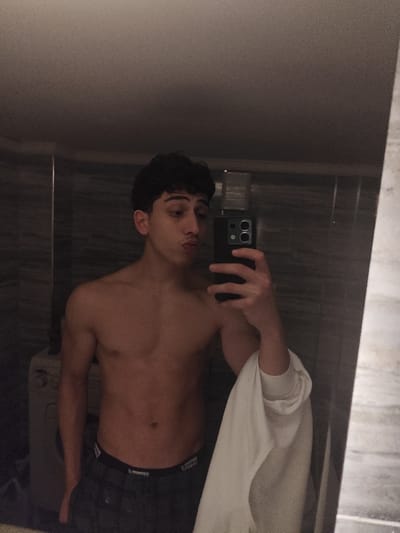 A shirtless young man with dark, curly hair pouts while taking a selfie in a bathroom mirror. He holds a black smartphone in his right hand, partially obscuring his face. He's wearing dark shorts and a white shirt is draped over his right arm. The bathroom has tiled walls and a washing machine is visible in the background. The lighting is somewhat dim, creating shadows on his body and the surrounding environment.