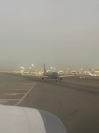 The image shows an aerial view of an airport tarmac at night or in low visibility conditions. Another airplane is visible on the tarmac, parked near a row of illuminated terminal buildings. The wing and engine of the aircraft the photograph was taken from are visible in the right foreground. Fog or haze obscures the background.
