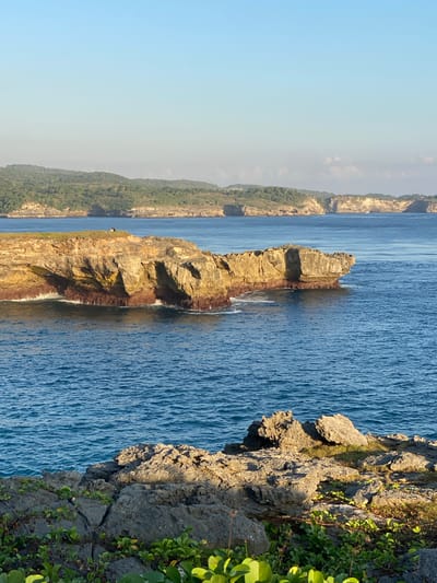 This media file displays a scenic view of the coastline in Nusa Penida, Indonesia. The image is oriented vertically, presenting a panoramic perspective of the ocean and the rugged cliffs.
The main subjects are the dramatic rock formations that jut out into the vibrant blue sea. The rocks are weathered and textured, showcasing layers of sedimentary rock. Some areas are covered in dark red algae near the waterline, indicating the influence of the tide. Lush green vegetation clings to the cliff faces in some areas, adding a touch of life to the otherwise stark landscape.
The scene is set along a coastline where the sea meets the land. In the distance, more hills covered in green foliage are visible, suggesting a larger island or mainland. The water is a deep, rich blue with a textured surface, indicating it is a relatively calm sea with gentle ripples.
There is no discernible activity or event taking place, it appears to be a still, natural landscape. A single, small person can be seen perched on a rocky outcrop in the mid-ground, providing a sense of scale to the immense geological features.
The time of day appears to be late afternoon or early evening, as the sun casts long shadows and highlights the textures of the rocks with a warm, golden light. The sky is a clear, pale blue, with a slight haze near the horizon. The overall mood is one of tranquility and the raw beauty of nature. No text is visible in the image.