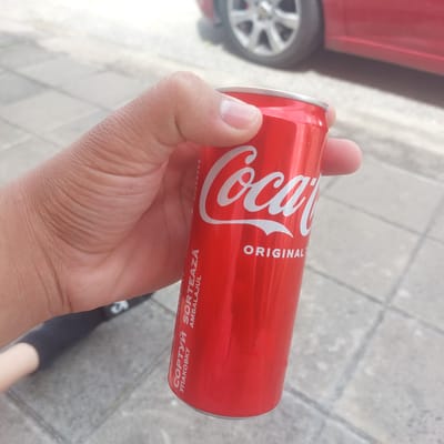 A person is holding a red Coca-Cola can in their right hand. The can has the Coca-Cola logo in white script and the word "ORIGINAL" below it. There are also some vertical lines of text in a foreign language on the left side of the can. The background is a blurred outdoor scene with a paved surface and a red car. The image appears to be taken in Ablanitsa, Bulgaria.