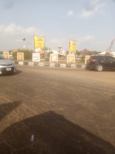 A sunny day in Unguwar Sarki, Nigeria, as a street scene unfolds with cars and advertising billboards. The foreground is dominated by a paved road with a shadow cast from the left, indicating the time of day is likely late morning or early afternoon. A silver sedan is parked on the left, its license plate visible as "OKA 2082". A black SUV is positioned further down the road on the right.

Behind the road, a central median features a series of advertising displays. The most prominent are large, vertical billboards, one of which clearly shows a yellow package with "Indomie" branding and an image of noodles with a fried egg. Another billboard shows a similar Indomie package. Other rectangular displays feature images of food and text, likely advertisements for various products. There's also a decorative fountain or sculpture in the background with a red archway. The scene appears to be a busy intersection or a commercial area. The sky is clear with some scattered clouds, suggesting fair weather. The overall impression is one of everyday life in a Nigerian urban setting, with commerce and transportation being central themes.