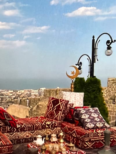 The image depicts an outdoor seating area overlooking a city and a body of water. The seating is arranged on a raised platform and features traditional Middle Eastern-style cushions and floor seating upholstered in rich red and patterned fabrics. Several decorative pillows are scattered on the seating. In the foreground, a table displays a white teapot and several ornate brass containers, possibly for serving drinks or sweets.
Behind the seating area, a stone wall forms part of the structure. A black street lamp with curved arms and two glass globes stands to the right, topped with a decorative crescent moon and star ornament. Two evergreen bushes are planted near the lamp.
In the background, a cityscape with buildings can be seen stretching towards the horizon, where it meets a vast expanse of water, likely a sea or large lake. The sky is blue with scattered white clouds. The architecture of the city appears to be a mix of older stone structures and more modern buildings. Two figures, one in white attire and another in darker clothing, are visible standing on a stepped stone area in the middle ground, overlooking the city. The overall impression is one of a scenic vantage point, possibly a cafe or a resting area in a historic location. The mention of Lopatino, Russia, implies the city is located in that region, though its specific appearance does not immediately suggest a Russian setting.