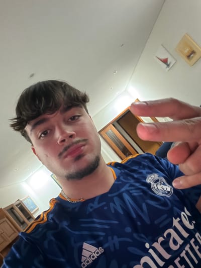 A young male subject is depicted in an indoor selfie, located in what is contextually identified as Gafanha da Nazaré, Portugal. The subject, appearing in his late teens or early twenties, has dark brown hair with a fringe and a light complexion. He displays a thin mustache and a goatee connected by subtle facial hair along the jawline. He is wearing a blue, short-sleeved Adidas t-shirt with orange trim on the shoulders and an abstract pattern of darker blue lines. The shirt features a white embroidered Real Madrid crest on the left chest and the "emirates" sponsor logo in white text. A silver-colored chain necklace is visible around his neck. His right hand is raised towards the camera, fingers extended in a two-finger gesture. The background shows light-colored walls and a ceiling fitted with multiple recessed light fixtures. Visible background elements include two framed pictures on the upper right wall, a closed wooden door with glass panels on the mid-right, and a section of wooden furniture on the lower left. The scene is brightly illuminated by internal light sources.
