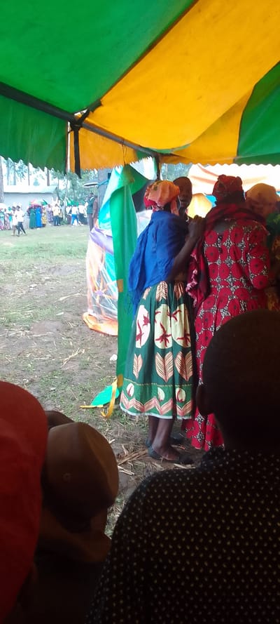 The image shows a scene inside a tent with a yellow and green striped canvas roof. Four individuals are gathered beneath the tent, the two closest to the camera are women. The woman on the left is wearing a blue shawl and a green and red patterned skirt, while the woman on the right is wearing a red and white patterned dress. The woman on the left is speaking to the woman on the right, who is listening. Behind them, a man wearing a black and white striped shirt is looking to his right. Behind the man, another woman wearing a red head scarf is partially visible. There is a large crowd of people outside the tent in the background. The ground is covered in short grass and dry stalks of plants. The scene is set in a rural location, potentially a village or town square in Kenya. The coordinates provided (latitude: 0.379, longitude: 34.78) correspond to a location in Lubao, Kenya.
