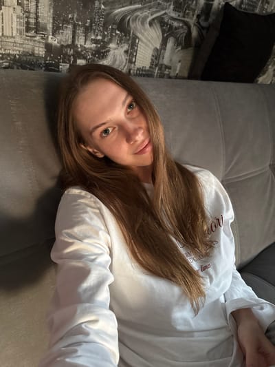 A young woman with long, brown hair and blue eyes is sitting on a gray couch, looking directly at the camera. She is wearing a white long-sleeved shirt with red text on it. The background features a black and white mural of a city skyline. The lighting suggests it is daytime, with a light source coming from the right. The woman appears to be relaxed and is leaning against the couch. There are no other people or animals visible in the image. The location is not specified within the image itself.