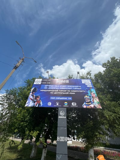 The image shows a billboard advertising an esports tournament in Kokshetau, Kazakhstan. The billboard, positioned against a blue sky with scattered clouds, features graphics of characters from a video game, likely representing participants or mascots. Text on the billboard is in both Russian and Kazakh, with details about the event such as dates ("5 июня, 12:00" and "5 июля, 12:00"), location ("МЕСТО: KOKSHE ARENA"), and event title ("ОРТАЛЫҚ АЗИЯ ЕЛДЕРІ АРАСЫНДАҒЫ ХАЛЫҚАРАЛЫҚ КИБЕРСПОРТ ТУРНИР ПО КИБЕРСПОРТУ ПО ЦЕНТРАЛЬНОЙ АЗИИ"). The address is given as "Мкр. Сарыарка, 17А". Below the main advertisement, logos for "Digital Aqmola" and "KAZAKHTELECOM" are visible. The billboard is mounted on a grey, rectangular pole with a decorative pattern near the bottom. In the background, lush green trees and a street are partially visible. A concrete utility pole with lights is also present in the frame, leaning to the left. The scene appears to be outdoors during daylight hours.
