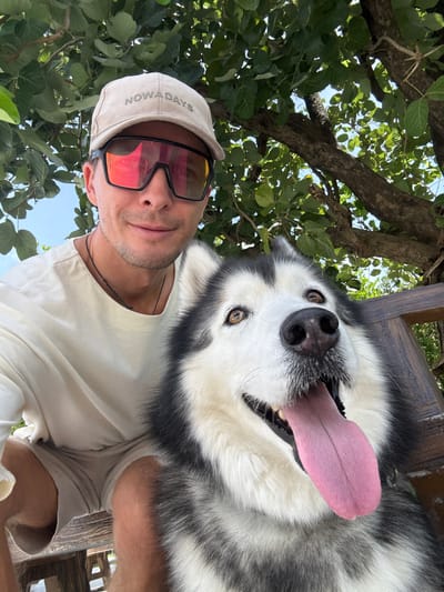 A man and a husky dog pose for a selfie outdoors, seated on a wooden bench under a leafy tree. The man, wearing a light-colored t-shirt, tan shorts, a beige baseball cap with "NOWADAYS" visible on it, and large, red-tinted sunglasses, smiles at the camera. The husky, with its mouth open and tongue out, looks directly at the camera with light brown eyes. The background features green foliage and blue sky, suggesting a warm, sunny day. The scene appears to be a relaxed, casual outdoor setting, possibly in Chalong, Thailand, given the presence of lush greenery and bright sunlight.