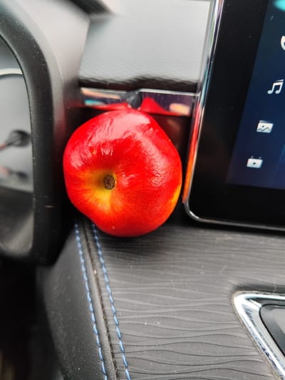 A close-up, slightly overhead shot shows a bright red apple with yellow accents resting on a textured black surface. The apple is positioned to the left of a dark rectangular screen, which displays icons resembling a musical note and a photograph. To the left of the apple, a portion of a black dashboard is visible. The background consists of the interior of a vehicle. This image was taken in Vsevolozhsk, Russia.