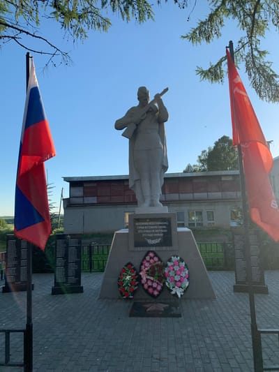 The image shows a war memorial in the city of Svyatitsa, Russia. In the foreground, two flags are displayed: the Russian flag on the left and a red flag, possibly a Soviet or communist flag, on the right. The central element of the memorial is a stone statue of a soldier holding a rifle. Below the statue is a monument with inscribed names and a quote. Wreaths of artificial flowers are placed at the base of the monument. The memorial is set on a paved area with a fence in the background. The environment appears to be outdoors with trees and a clear blue sky. Buildings are visible behind the memorial.