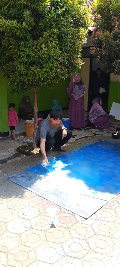 A young man, wearing a grey t-shirt and black pants, is squatting on a paved surface in front of a large blue tarp. He is holding a blue spray can in his right hand and spraying it onto the tarp, which is lying on the ground. The tarp is already partially painted blue. There is a large green tree behind him and a paved area of hexagonal tiles in front of him. The man is facing the viewer. There are five other people in the background, including two women in purple dresses, and a small girl in a pink dress, standing by the tree. They are all looking at the man. The location is Cangkuang, Indonesia. The coordinates are -7.065 latitude and 107.552 longitude. There are two buckets, one yellow and one blue, sitting on the ground, in front of the tree. A building can be seen to the right of the tree.