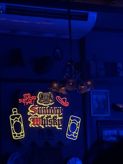 The image depicts a dimly lit interior, possibly a bar or restaurant, bathed in blue and purple light. Dominating the scene is a neon sign that reads "IT'S SUNTORY TIME!" in red lettering, with "Suntory Whisky" written below in yellow. Next to the sign, on the left, is a neon outline of a bottle, and on the right, another neon outline of a bottle with a circular design within. Above the neon sign, a chandelier with multiple illuminated flower-like shades hangs from a chain. Further up, an air conditioning unit is visible on the ceiling. The background features dark walls with framed pictures and some shadowed objects. There are no visible people in the immediate foreground or midground. The overall ambiance is moody and atmospheric, with a focus on the glowing neon signage. The image is taken from a low angle, looking upwards.