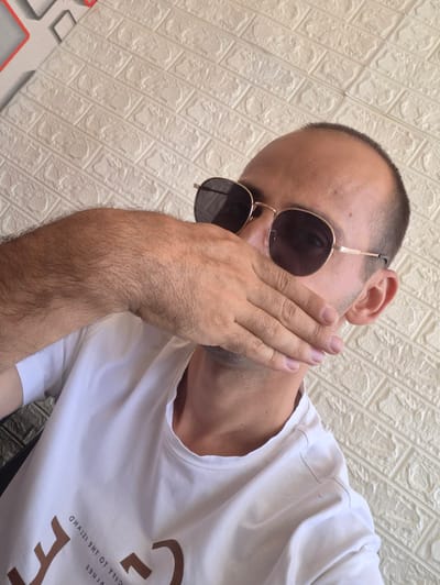 A man is captured in a close-up, selfie-style shot, wearing sunglasses and a white t-shirt. His right arm is raised, covering his mouth and chin. The background is a textured, white brick-like wall. The man has a shaved head and visible stubble. The t-shirt has a graphic design on the lower front, though partially obscured. The image composition suggests a casual setting, potentially indoors.