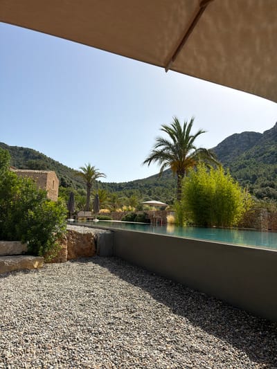 The image depicts an outdoor scene at a resort or private residence in Artà, Spain, featuring a contemporary swimming pool and surrounding landscape. A large, beige umbrella partially obstructs the top of the frame, casting a shadow over the sky and upper portion of the view.

The foreground is dominated by a gravel pathway leading towards the pool. To the left, large natural stones and lush green bushes are visible, with a rustic stone building in the background. Two grey umbrellas are positioned near the pool's edge, along with lounge chairs and a small table, suggesting a seating area.

The swimming pool itself is elongated and appears to have an infinity edge, with clear turquoise water. Beyond the pool, the landscape transitions into a lush, green environment with palm trees and dense vegetation. In the distance, a majestic mountain range covered in trees rises against a clear blue sky. The architecture of the stone building and the natural elements of the landscape suggest a Mediterranean setting.