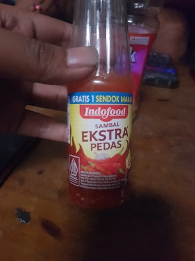 The scene, located indoors in Cimenyan, Indonesia, features a human hand holding a clear glass bottle of Indofood Sambal Ekstra Pedas chili sauce. The bottle contains a red, viscous liquid. Its yellow and blue label displays the brand "Indofood," the product name "SAMBAL EKSTRA PEDAS," the text "GRATIS 1 SENDOK MAKAN," and a Halal certification logo. Illustrations of red chili peppers and garlic are also present on the label. The hand, positioned on the left, shows a single visible fingernail and grips the bottle firmly. The immediate foreground consists of a wooden surface, exhibiting visible grain and minor blemishes. The background is dimly lit and out of focus, revealing a partial view of a dark pink or red container and an unidentified dark rectangular object.
