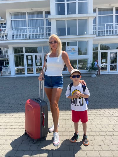 A woman and a young boy stand on a paved walkway in front of a multi-story white building, likely a hotel, in Giachrypsh, Abkhazia. The woman, with blonde hair and sunglasses, wears a white tank top and denim shorts. She holds the handle of a red suitcase. The boy, wearing a white t-shirt, red shorts, a cap, and sunglasses, stands next to her with his hands clasped. The building features large windows and balconies. The walkway is paved with grey rectangular tiles. Sunlight casts shadows on the ground.