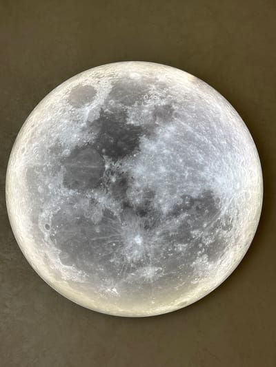This media file features a close-up, illuminated replica of the moon against a textured, dark brown background. The moon's surface is detailed with craters of varying sizes and shades of gray, interspersed with brighter, more reflective areas, suggesting a representation of its actual topography. The light source appears to be internal, casting a soft glow and highlighting the three-dimensional texture of the lunar surface. The overall impression is one of a decorative object or display rather than a natural celestial body. There is no discernible activity or event taking place, and no visible text. The setting is abstract, with the dark background providing a neutral contrast to the bright moon, making it the sole focal point. The scene is static and appears to be indoors, given the controlled lighting and the nature of the object depicted.