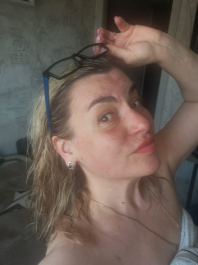 A woman is posing in a selfie-style photograph. She is fair-skinned with wet, blonde hair. She wears a gold chain necklace and an earring in her left ear. Glasses with black frames and blue temples are resting on her forehead. Her left hand is raised near her head, and she is wearing a ring on her ring finger. The background appears to be a wall and possibly other furniture or objects that are out of focus. There is no information in the image to determine that it was taken in Elektrostal, Russia.