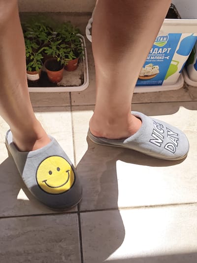 A person's legs and feet, wearing gray slippers, are visible in the image. One slipper has a yellow smiley face design, and the other has "NICE DAY" embroidered on it. The person is standing on a tiled floor, possibly outdoors, as evidenced by the sunlight casting shadows. In the background, a white container with blue lettering, which appears to be a food product, and a tray of young tomato plants in small pots are present. The overall scene suggests a casual, everyday moment. The context of Pazardzhik, Bulgaria, suggests this might be a domestic setting or a garden area.