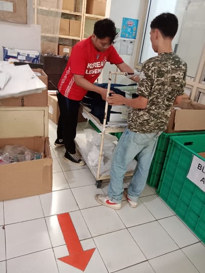 Two people are working in a warehouse. One person wears a red shirt with "KOREA LEGEND" printed on it and black pants, while the other person wears a camouflage shirt and jeans. They are working together to move items on a metal cart. Cardboard boxes and green crates surround them. An orange arrow is on the tiled floor, pointing away from the people.
