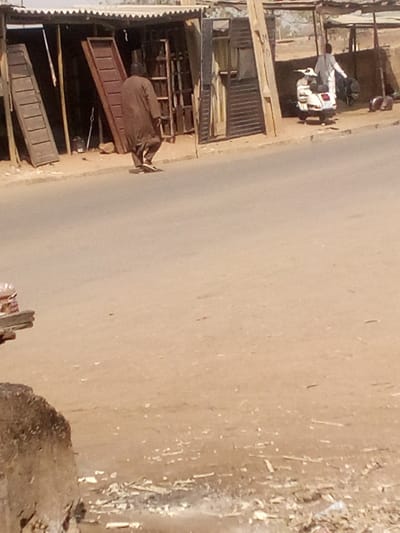 This image depicts a daytime street scene in Tudun Murtala, Nigeria. The foreground shows a dusty, light brown dirt shoulder with scattered white debris. A paved, grey-brown road runs horizontally across the midground.
In the background, on the left, a man wearing a long, dark brown traditional garment and dark headwear is walking away from the camera, moving towards the right side of the frame. He wears dark open-toed sandals. Behind him is an open-fronted workshop or stall with a corrugated metal roof supported by wooden poles. Inside and around this structure, multiple brown wooden door panels or shutters are visible, some leaning against walls, indicating a carpentry or door-making business.
Further to the right in the background, another man in a light-colored long-sleeved top and trousers is standing near a white scooter, possibly a Vespa, which is parked on the dirt shoulder. The man appears to be facing or attending to the scooter or an object behind it. Behind him, additional rudimentary structures with roofs are visible. Two light-brown animals, possibly goats, are on the ground near these structures on the far right. The overall environment suggests a roadside commercial area.