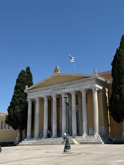 The image shows a bright, sunny day in Athens, Greece. A grand neoclassical building with a prominent portico of Ionic columns dominates the scene. The building is made of marble and painted in a pale yellow. The name "ΖΑΠΠΕΙΟΝ" (Zappeion) is inscribed in Greek capital letters on the entablature above the columns. A Greek flag, blue and white, flies from a flagpole to the left of the building. Tall, dark green cypress trees stand beside the building, providing a stark contrast to the pale facade and the clear blue sky.
Several people are visible in the image. In the foreground, a person is standing near a lamppost in front of the building's steps. Further up the steps, a couple of individuals are seen walking or standing. On a higher level of the building, another person is leaning out of an opening. The overall atmosphere suggests a tourist destination on a pleasant day, with visitors exploring the historic site. The sunlight casts sharp shadows, indicating it is likely midday. The architecture and the Greek flag clearly identify the location as Greece, specifically the Zappeion Hall, a prominent landmark in Athens often used for events and public gatherings.