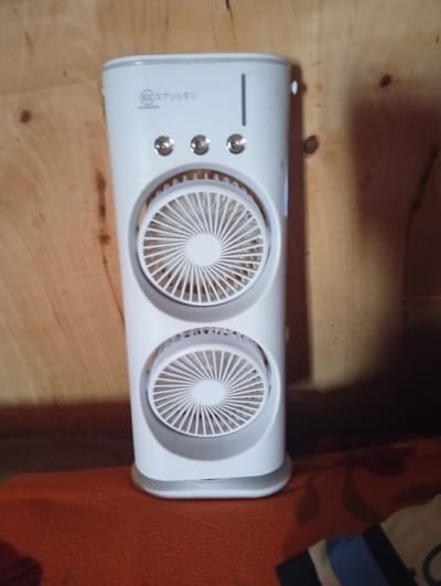The image shows a white, tower-style personal fan with two circular fan grills. The fan is positioned on what appears to be an orange surface, possibly a piece of furniture or bedding. The background is a textured wooden wall. The fan has two control buttons on the top section and a vertical slit indicating the water level. The model name "SX スアンシオン" can be seen at the top of the fan. There is no evidence of Igrita, Nigeria or any other identifiable location. There are no people or actions visible.