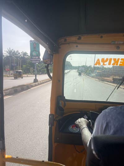 The image provides a forward-facing perspective from inside a yellow Keke Napep (tricycle taxi) on a sunny day in Barnawa, Nigeria. The driver's right arm, wearing a silver-colored bracelet, is visible on the steering handlebar. The Keke's windshield exhibits significant damage, with a prominent spiderweb fracture originating from the lower right quadrant and multiple linear cracks extending across the glass. The word "AYIKE" is visible in reversed orange lettering, appearing as "EKIYA," on the upper right interior of the windshield.

Outside the Keke, a wide, paved road with a central median is visible, featuring white dashed lane markings. Another yellow Keke Napep is observed traveling ahead in the same direction on the left side of the road. Further along the left roadside, a tall billboard displays a man's face on a green background, with the partially visible text "Opay," a large "N" symbol, and the phrase "Large Transaction Shield." Palm trees and other vegetation are visible behind the billboard. In the distance, a dark-colored vehicle is also proceeding along the road. The right side of the road reveals an undeveloped or construction area with piles of aggregate and concrete blocks. The sky above is clear.