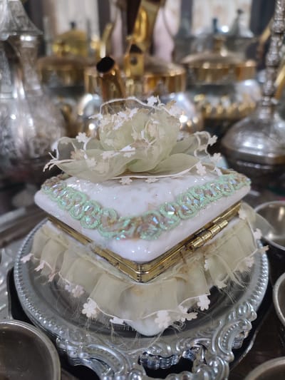 The image shows a close-up of a decorative white and green trinket box with floral accents, resting on a silver tray. The background is blurred, featuring what appear to be ornate metal teapots and other decorative items, possibly in a display or shop setting. There are no people or animals visible in the image. The location in Blang Cut, Indonesia, is not discernible from the visual information presented in the photograph.
