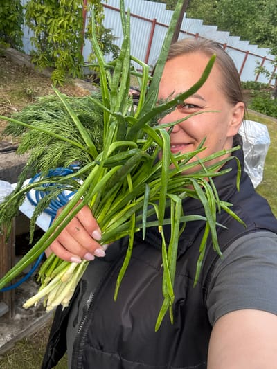 A woman is holding a bunch of green onions and dill in her hand, smiling. She is wearing a gray shirt and a black vest. The background is a garden with a metal fence and some greenery. The location is inferred to be Dmitrov, Russia, but this is not verifiable from the image alone.