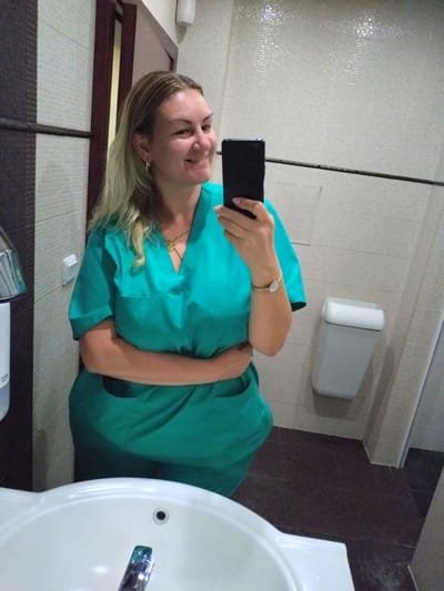 A woman is taking a mirror selfie in a bathroom. She is wearing a green scrub suit and has blonde hair. She is smiling and holding a black smartphone in her right hand. Her left arm is crossed over her chest. She is wearing a gold necklace and a gold wristwatch. The bathroom has tiled walls and floor. A white sink and a toilet paper dispenser are visible. The image is undated and no location information is available.