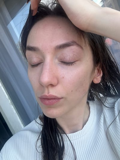 A close-up, slightly above eye-level shot shows a young woman's face with her eyes closed. She has dark, wet hair that is partially pulled back by her right hand, which is resting on the top of her head. The woman is wearing a white, ribbed top. Her skin appears fair with some freckles and minor blemishes. The background consists of a window with vertical blinds, suggesting an indoor setting. No specific location in Vracar Urban Municipality, Serbia can be determined from the image.