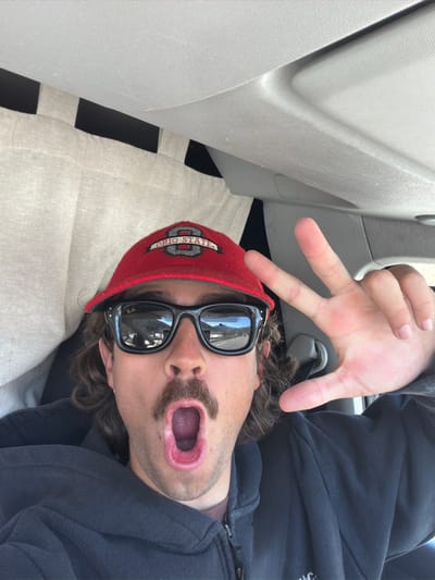A male individual is photographed in a close-up, high-angle view from inside a vehicle in Saint-Chaffrey, France. The subject wears a red "Ohio State" baseball cap and black Wayfarer-style sunglasses. He has a dark mustache and long wavy brown hair. His mouth is wide open, revealing teeth and tongue. He wears a dark zippered hooded sweatshirt over a lighter brown shirt. His left hand is raised, displaying extended index and middle fingers, with the ring and pinky fingers curled, and the thumb bent towards the palm. The reflections in the sunglasses show a road, a guardrail, and mountains under a clear blue sky. The vehicle's interior includes light-colored fabric paneling on the left and a light grey ceiling.
