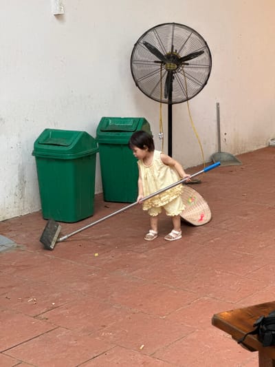 A young child in a light yellow dress and sandals is sweeping a reddish-brown tiled outdoor area with a long-handled broom. Behind the child are two large green trash cans and a large industrial fan mounted on a stand. The wall behind the objects is a rough, off-white texture. A woven basket or hat is lying on the ground near the child. The scene appears to be in an outdoor, possibly covered, area with a concrete floor. The general ambiance suggests a casual, everyday setting, perhaps in a residential or communal space.