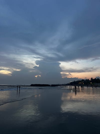 The image shows a beach scene at sunset in Canggu, Indonesia. The sky is filled with clouds, varying in color from dark blue to orange. The ocean reflects the colors of the sky. Silhouettes of people can be seen walking along the beach. There are buildings and trees in the distance.