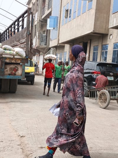 The image captures an urban street scene in Fagge C, Nigeria, during daylight. In the foreground, a woman walks from right to left, observed from her left side. She wears a long, patterned dress in maroon, black, and white, topped with a dark blue hijab. Her left hand holds multiple white papers, while her right hand grips a light-colored smartphone. Her left lower leg is encased in a dark cast or brace, adorned with colorful fabric elements around the ankle.
In the middle ground, a man in a red t-shirt and dark shorts walks away from the viewer, balancing a large white sack on his head. To his right and slightly behind him, several other men are visible; at least three wear green t-shirts, with some wearing white caps.
To the far left, the rear section of a large, blue utility truck is present, its bed loaded with multiple white sacks of goods. A yellow compact utility vehicle is partially visible behind the truck. On the right side of the frame, a black SUV with a "LAGOS TYU 964HY" license plate is parked. Behind the SUV, a man in a red and black patterned shirt sits on a wooden handcart featuring large, spoked wheels.
The background consists of multi-story buildings; some display modern blue-tinted glass windows, while others exhibit exposed concrete and rebar, suggesting ongoing construction or various states of repair. The ground is a light-colored, paved surface. A metal structure providing overhead cover is partially visible in the upper left.