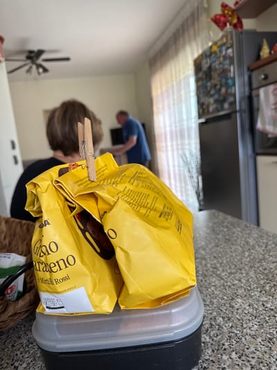 The image shows a partially opened, yellow bag of "Golosina Caraxeno Mirtilli Rossi" cookies secured with a clothespin. The bag sits atop a clear plastic container with a black lid. In the background, out of focus, there are two people. A man in a blue shirt is standing. Behind the bag is a woman with brown hair. The image appears to be taken indoors in a kitchen setting. Based on the product name and labeling (Mirtilli Rossi), this food product is likely from Italy, and Aprilia, Italy is not directly identifiable in the image or labeling, but that doesn't exclude Aprilia as the location.