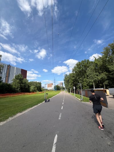 A man in a black t-shirt and black shorts is doing push-ups on an asphalt path in a park. He is wearing red and white sneakers. The path has white dashed lines painted on it, indicating a lane. To the left of the path is a strip of green grass, and beyond that are trees and power lines. In the background, there are several tall residential buildings. A scooter is parked on the grass near the path. The sky is blue with scattered white clouds, suggesting it is a sunny day. The lighting and shadows indicate it is daytime. The overall scene appears to be a public park area in a city, likely Moskovskiy, Russia, given the building architecture. There is no visible text in the image.