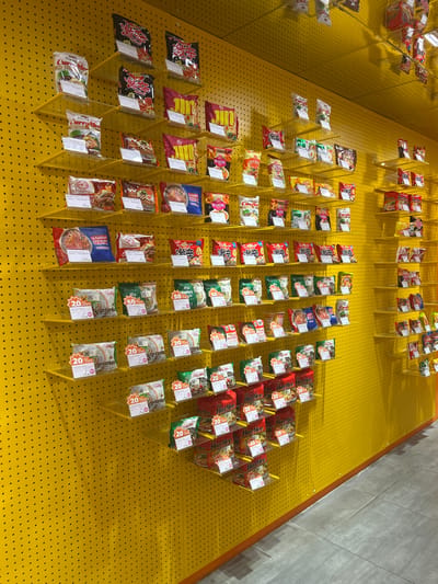 The image shows a retail display of instant noodles. The noodles are arranged on a yellow, perforated wall with clear plastic holders. The display is sectioned into rows and columns, with multiple varieties of noodles presented. Some of the packages are red, black, or white, and many have visible text in Asian languages. Labels on some packages indicate prices and discounts, such as "20% off". A prominent brand displayed is "Harlee". The setting appears to be indoors, likely a market or a store, given the organized retail display and the presence of a concrete wall in the background. The lighting is bright and even, suggesting artificial light. The overall scene is clean and organized, with no people visible. The context provided indicates the location is Makassar, Indonesia.