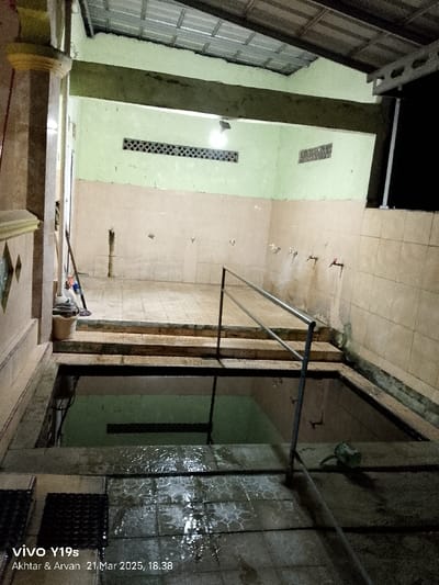 The image shows the inside of a tiled, rectangular room. The room has a green tiled floor and a tiled wall on the right side of the image. On the opposite side of the wall is a black metal railing. The railing goes across a short set of stairs leading to a concrete area. The concrete area has more tiled walls, including a white and green wall on the far side of the room. There are several visible faucets on the walls, and the area is mostly empty. Behind the railing, the concrete area continues down to a large, rectangular, open pool filled with green water. The pool has a visible tiled floor. The concrete area is wet and has a drainage system, visible in the lower-right corner of the image. The room is dimly lit, likely lit by a single, overhead, bright light fixture on the ceiling. The location is a public bathhouse, possibly attached to a mosque, in the Kecamatan Kroya area of Indonesia. The latitude and longitude coordinates provided are for a location in Kecamatan Kroya.