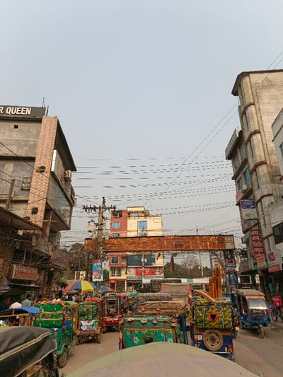This image depicts a densely populated urban street scene in Savar, Bangladesh, under a clear or hazy daytime sky. The street is heavily congested with a variety of vehicles, primarily numerous three-wheeled auto-rickshaws adorned with colorful tarpaulins and fabrics, including green, blue, and camouflage patterns.
Multi-story commercial and residential buildings line both sides of the street. On the left, a grey concrete building with "SUPER QUEEN" visible at its upper level features a section clad in light brown brick-like material. On the right, another multi-story building, light brown in hue, exhibits a somewhat rounded corner design and multiple window frames. Between these main structures, in the background, several other buildings are visible, including one with prominent red and light blue sections, and another below it displaying "FURNITURE Concept" signage.
Above the street, an extensive and dense network of overhead utility wires crisscrosses, connected to multiple utility poles equipped with transformers. A prominent, large, rusty metal structure, resembling an old overhead gantry or sign support, spans across the street in the mid-ground.
Numerous people are present, including pedestrians on the sidewalks, individuals seemingly engaged in commercial activities near vendor stalls on the left side, and drivers/passengers within the stationary or slow-moving auto-rickshaws. One individual is visible under a yellow and blue umbrella in the mid-left. Commercial signage, predominantly in Bengali script, is affixed to building facades and utility poles throughout the scene.