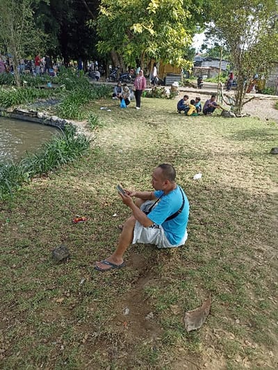 This image captures a vibrant outdoor scene, likely a park or recreational area in Mataram, Indonesia, during daytime. The weather appears pleasant, with dappled sunlight suggesting either late morning or afternoon.
In the immediate foreground, a man is seated on the grass, wearing a blue t-shirt, white shorts, and dark flip-flops. He is holding a smartphone in both hands and appears to be engrossed in it. A black strap from a bag is visible across his right shoulder. To his left, on the grass, a discarded red and white snack wrapper is visible.
To the left of the main subject, a tranquil body of water, possibly a pond or canal, stretches across the mid-ground, bordered by dense green aquatic plants and rocks. Beyond this water feature, and further into the background, numerous people are scattered across the grassy area under the shade of large, leafy trees.
In the mid-ground, a woman wearing a pink top and a light-colored headscarf is standing, looking towards the left. Nearby, a group of children and adults are squatting or sitting on the grass, some appearing to be observing something on the ground. Other individuals and small groups are visible throughout the background, engaged in various activities like sitting, standing, and strolling, contributing to a relaxed and social atmosphere.
The scene suggests a public gathering or leisure activity, with people enjoying the green space and each other's company. No discernible text is clearly visible on any objects or clothing.