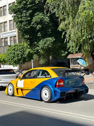In Gorna Oryahovitsa, Bulgaria, a custom-modified, bright yellow and blue Opel Astra race car is parked on an asphalt street. The car features a prominent rear spoiler, aerodynamic body kit, and white racing wheels. Next to the car, a white sedan is partially visible. In the background, a multi-story building with numerous windows stands behind lush green trees, and a small kiosk with a blue umbrella is also present. The sky is clear and the lighting suggests a sunny day.