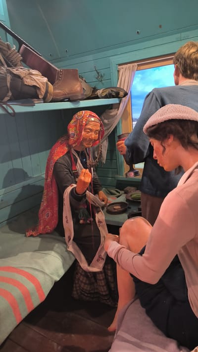 This image depicts a diorama inside a museum or exhibition, set within what appears to be the interior of a train carriage. The scene is populated by mannequins posed to represent a historical or cultural tableau.

In the foreground, a young woman wearing a light-colored cardigan and a white cap is seated, looking down. She has her right hand extended, seemingly interacting with the elderly woman in front of her. Her legs are crossed, and she is covered in a grey and white striped blanket.

The central figure is an elderly woman, seated and dressed in traditional clothing. She wears a red, patterned headscarf and a dark outer garment over a plaid skirt. She has a necklace of coins and is actively engaged in what appears to be sewing or crafting, holding a needle and thread. A white scarf with embroidered details is draped around her.

To the right, a third mannequin, a man, is partially visible, wearing a blue sweater and holding a cup. He is looking out of a window.

The environment is enclosed, with teal-colored wooden paneling on the walls and ceiling. A shelf above contains a brown boot, a rolled-up rope, and a guitar case, suggesting travel. A curtained window reveals a backdrop of a winter landscape with a blue sky and snow-covered trees. The floor is made of dark wooden planks.

The overall impression is that of a staged scene designed to evoke a sense of daily life or historical narrative from a specific cultural context. The location in Homyel, Belarus, is not directly evident from the visual cues within the image itself.