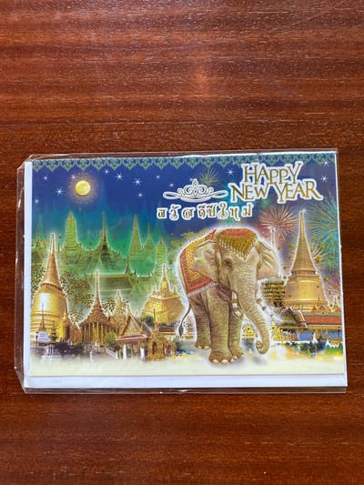 The image displays a New Year's greeting card featuring a scenic depiction of a Thai temple complex, possibly the Grand Palace in Bangkok, against a dark blue, starry night sky illuminated by fireworks. A majestic elephant, adorned with decorative coverings, stands prominently in the foreground, facing right. The card also includes the text "HAPPY NEW YEAR" in English and Thai script. The card is enclosed in a clear plastic sleeve and rests on a polished wooden surface, likely a table. The lighting suggests it is indoors, with the wooden surface exhibiting visible grain and reflections. There is no identifiable information suggesting the location is Bang Sare, Thailand.