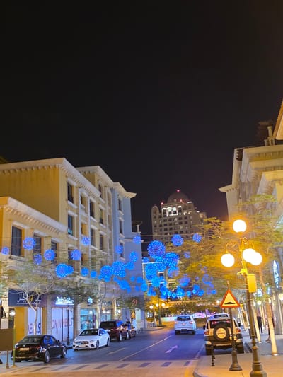 The image captures a nighttime street scene in Doha, Qatar, likely within the Katara Cultural Village, known for its distinctive architecture and vibrant atmosphere. The street is lined with buildings featuring traditional Qatari design elements, such as cream-colored facades and ornate detailing, illuminated by decorative blue lights strung across the street and from some of the building exteriors. Tall, modern buildings, one with a prominent domed roof, are visible in the background, contrasting with the more traditional architecture. Several vehicles, including a black sedan, a white sedan, a dark SUV, and a silver SUV with a spare tire mounted on the back, are parked or driving on the street. Trees with yellowing leaves are interspersed along the sidewalks, adding a touch of nature to the urban landscape. A streetlamp with multiple glowing orbs provides warm illumination, and a traffic sign warning of a speed bump is visible. A few pedestrians are seen walking along the sidewalk, contributing to the sense of an active urban environment. The overall impression is one of a festive and well-lit urban setting, characteristic of popular tourist and cultural destinations in Doha.