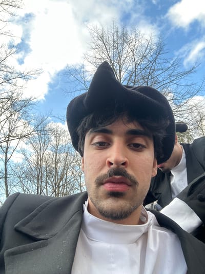 The image depicts an outdoor scene in Guimarães, Portugal, on a day with a partly cloudy blue sky and numerous leafless deciduous trees. The primary subject is an adult male, likely young, with dark hair, a prominent dark mustache, and a short beard. He is looking directly forward, possibly at the camera. He wears a black, felt-like hat with a distinctively folded or peaked crown, a black overcoat or jacket with lapels, and a white, high-collared shirt. Partially visible behind and to the right of the primary subject is another individual, also appearing male, wearing similar dark attire including a black coat and a white shirt, with a dark hat visible on their head. Both individuals are positioned within a natural environment characterized by the bare trees, suggesting a winter or early spring setting.